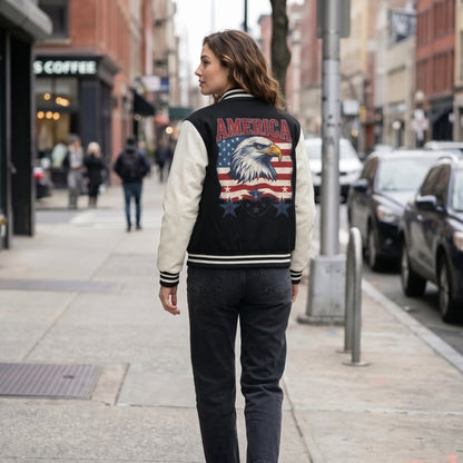 A woman walking on a city street wearing a black and cream varsity-style jacket with a large American flag and bald eagle graphic on the back, paired with dark jeans.