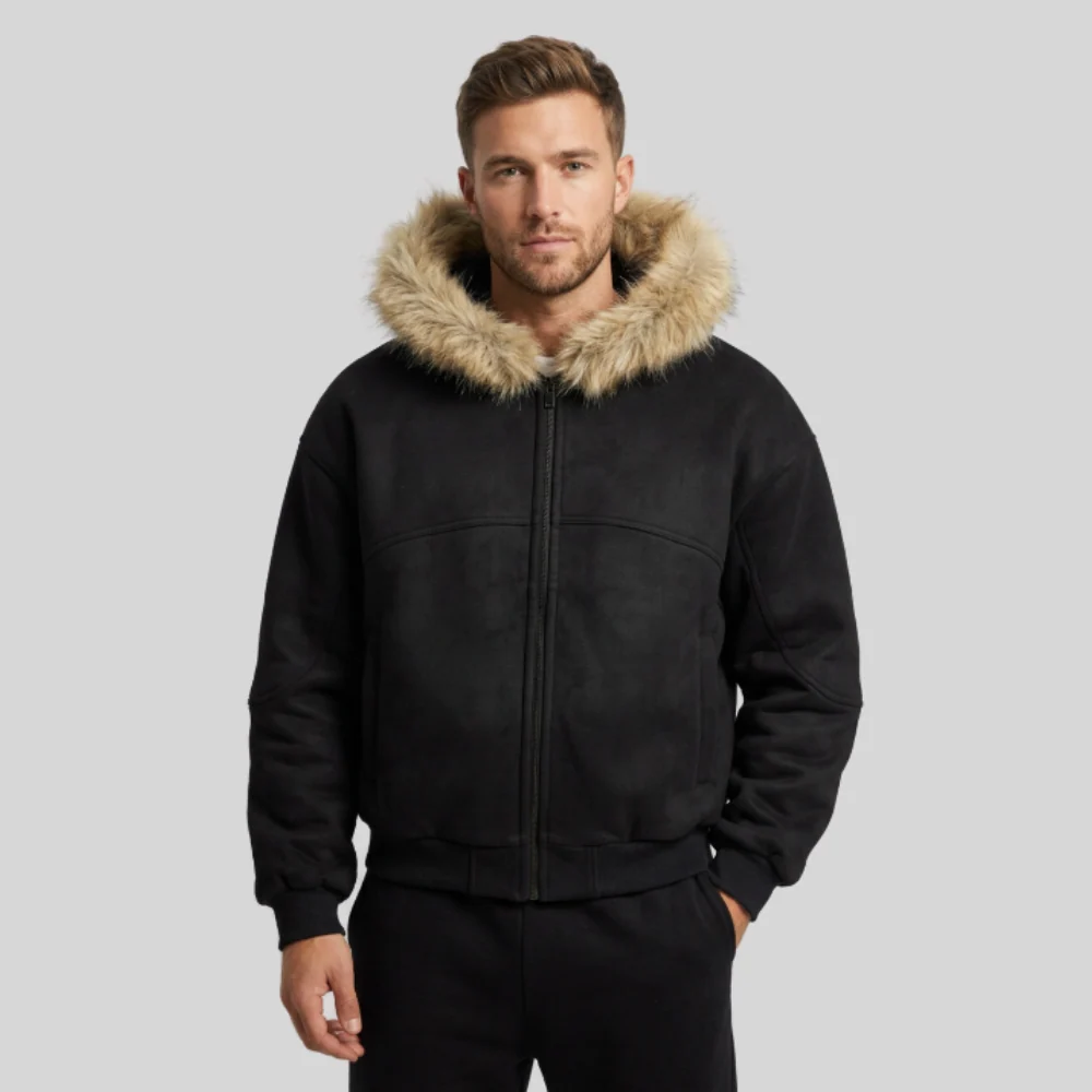 A man wearing a black zip-up jacket with a beige faux-fur trimmed hood, paired with black pants, photographed against a solid light gray studio background.
