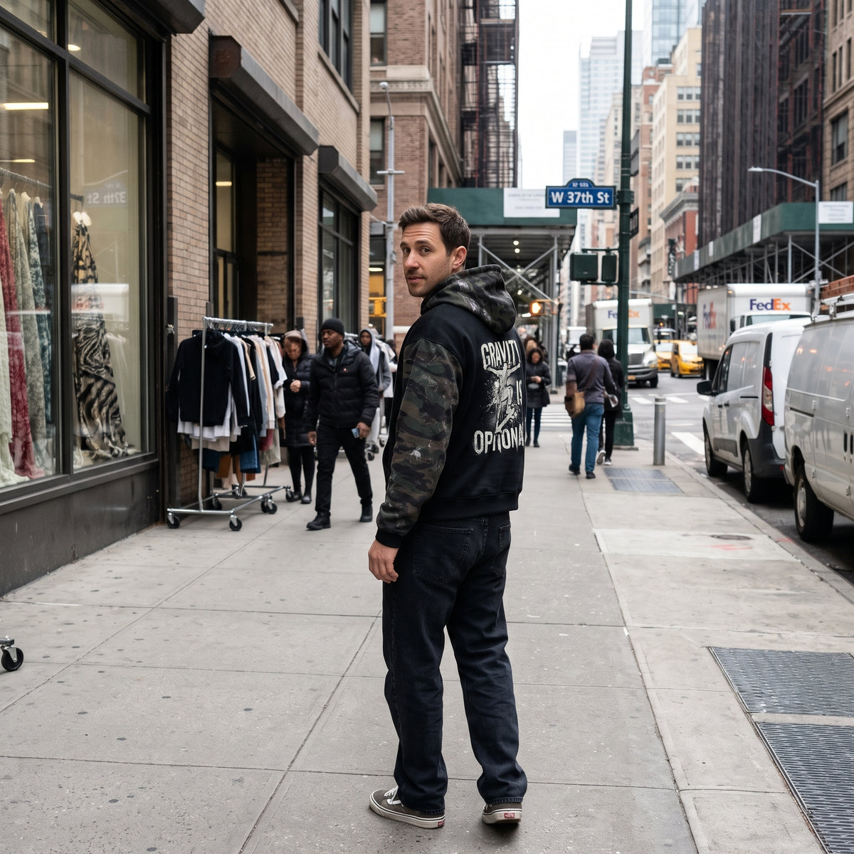 A man stands on a city sidewalk looking back over his shoulder. He is wearing a black hoodie with 'GRAND OPTION' printed in white on the back, dark jeans, and black sneakers.