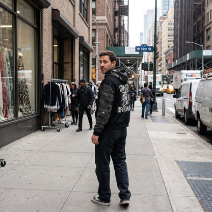 A man stands on a city sidewalk looking back over his shoulder. He is wearing a black hoodie with 'GRAND OPTION' printed in white on the back, dark jeans, and black sneakers.