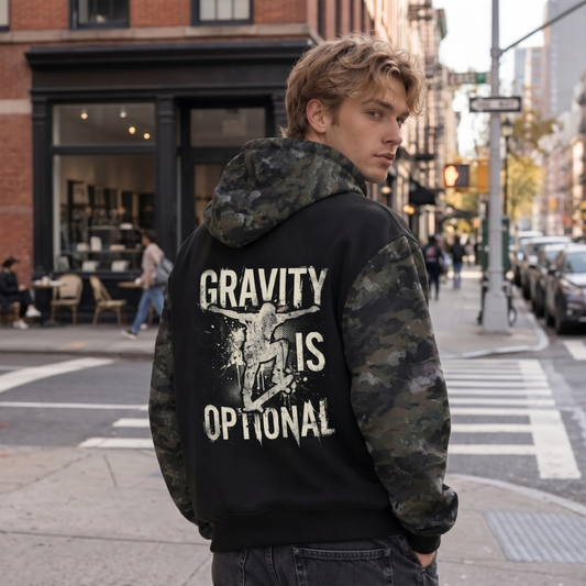 A man standing on a city street wearing a black hoodie with camouflage sleeves and hood, featuring the text 'GRAVITY IS OPTIONAL' in distressed white lettering on the back.